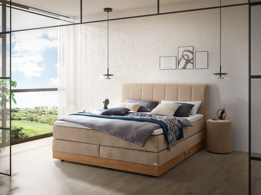 Ein helles Schlafzimmer mit einem beigefarbenen Boxspringbett mit vertikal gestepptem Kopfteil und einem Holzbettrahmen.