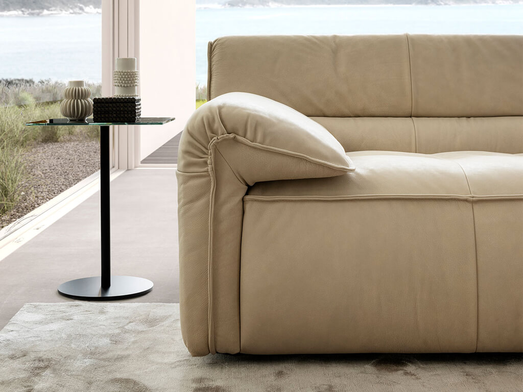 Nahaufnahme der Armlehne und des Rückens eines cremefarbenen Ledersofas, mit einem modernen Beistelltisch mit Vasen im Vordergrund und einem Fensterblick auf ein Gewässer und Hügel im Hintergrund.