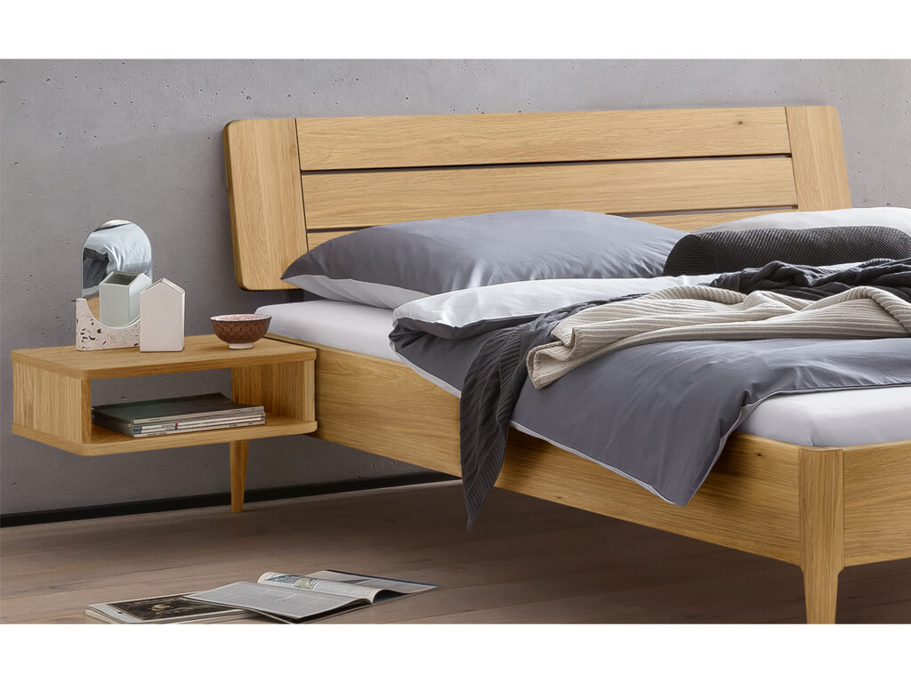 Eine Nahaufnahme des Kopfendes des Bettes "Busseto" aus heller Eiche, das ein Holzkopfteil mit horizontalen Lamellen aufweist. Ein passender Nachttisch ist am Bett befestigt.