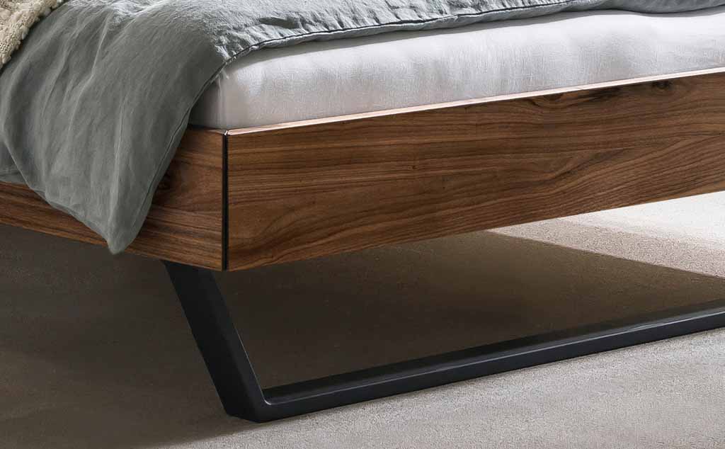 Nahaufnahme eines modernen Bettgestells aus Holz mit dunklen Metallfüßen, grauem Bettbezug und heller Matratze