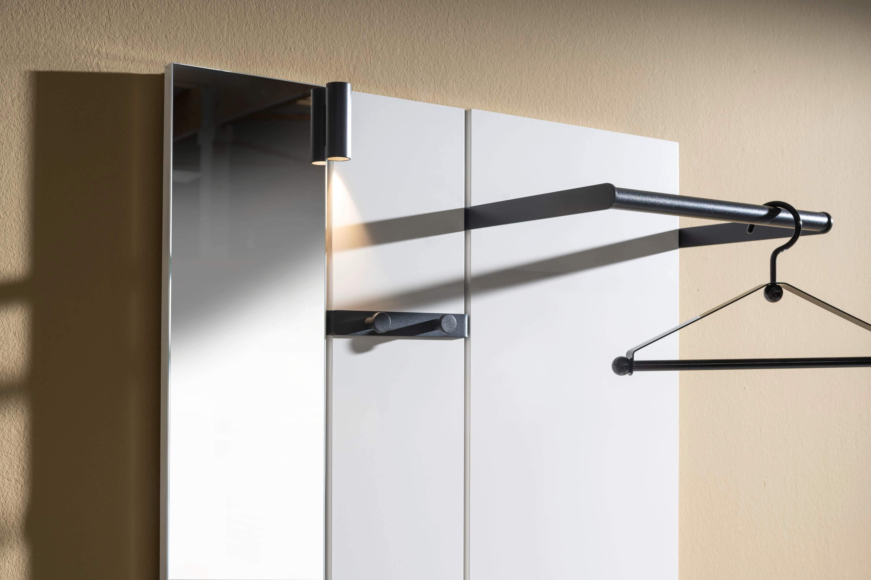Detailaufnahme eines modernen Garderobenspiegels mit integrierter Beleuchtung und einem schwarzen Kleiderbügel an einer Wand.