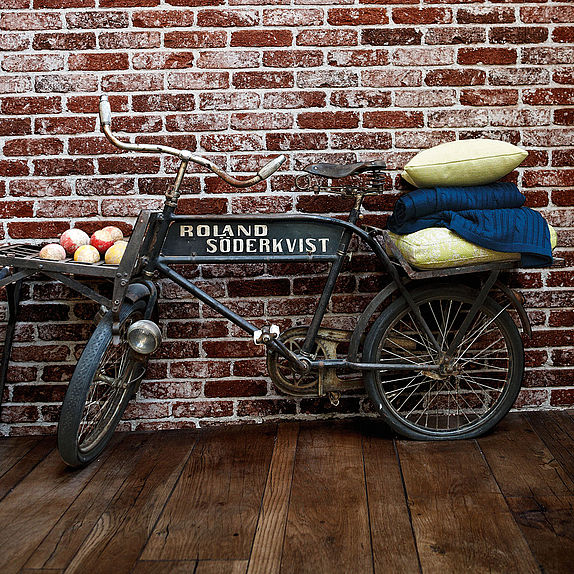 Ein altes Lastenfahrrad mit der Aufschrift 'Roland Söderkvist' steht vor einer Ziegelwand. Auf dem Fahrrad liegen Kissen und Decken, während im Transportkorb Äpfel liegen.