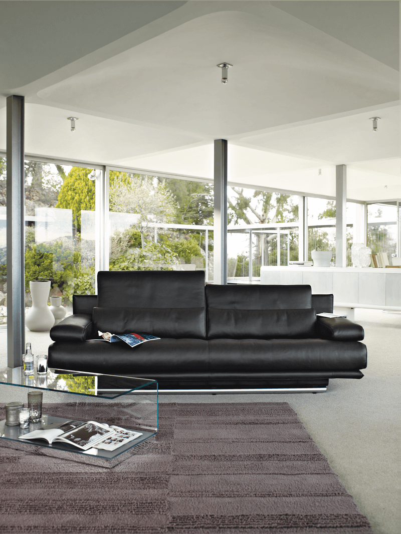 Schwarzes Ledersofa und Glastisch in einem hellen Wohnzimmer mit großen Fenstern