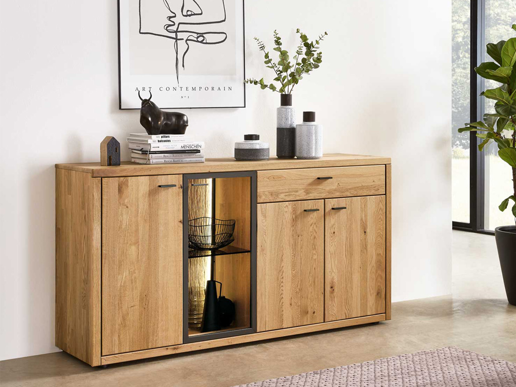Wöstmann WM 2250 - Sideboard | Type 2673/2674 | Wildeiche massiv | B: 168,0 cm Eleganter Holzschrank mit Glasvitrine, dekoriert mit Büchern, Kerzen und Pflanzen, vor einer weißen Wand mit einem Kunstplakat
