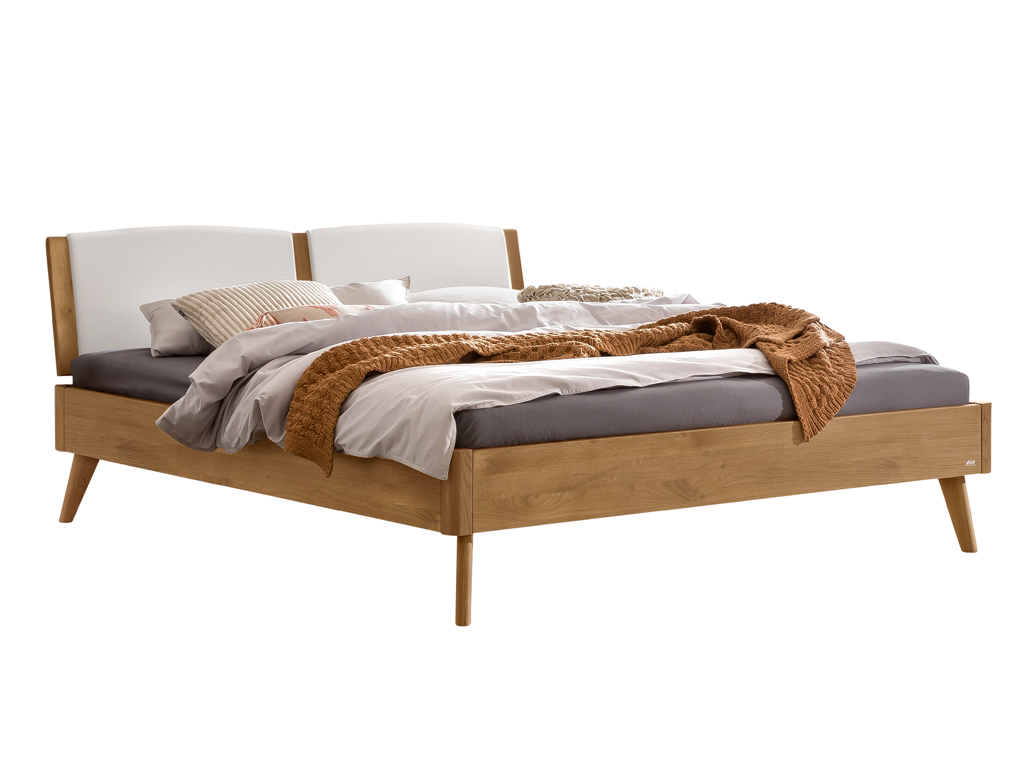 Ein modernes Doppelbett mit einem Gestell aus natur geölter Wildeiche und einem geteilten, weißen, gepolsterten Kopfteil. Es ist mit einer braunen Strickdecke und neutralen Bettwaren dekoriert. Die Holzmaserung des Gestells ist gut zu erkennen.