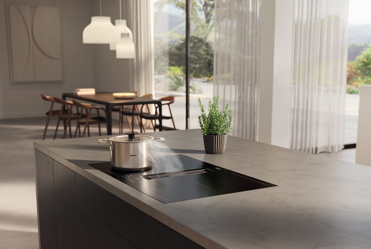 Ein Miele Kochfeld mit integriertem Dunstabzug in einer modernen Küche. Auf dem Kochfeld steht ein Edelstahltopf, aus dem Dampf aufsteigt. Eine kleine Topfpflanze steht daneben. Der Kochbereich ist Teil einer großen Kücheninsel.