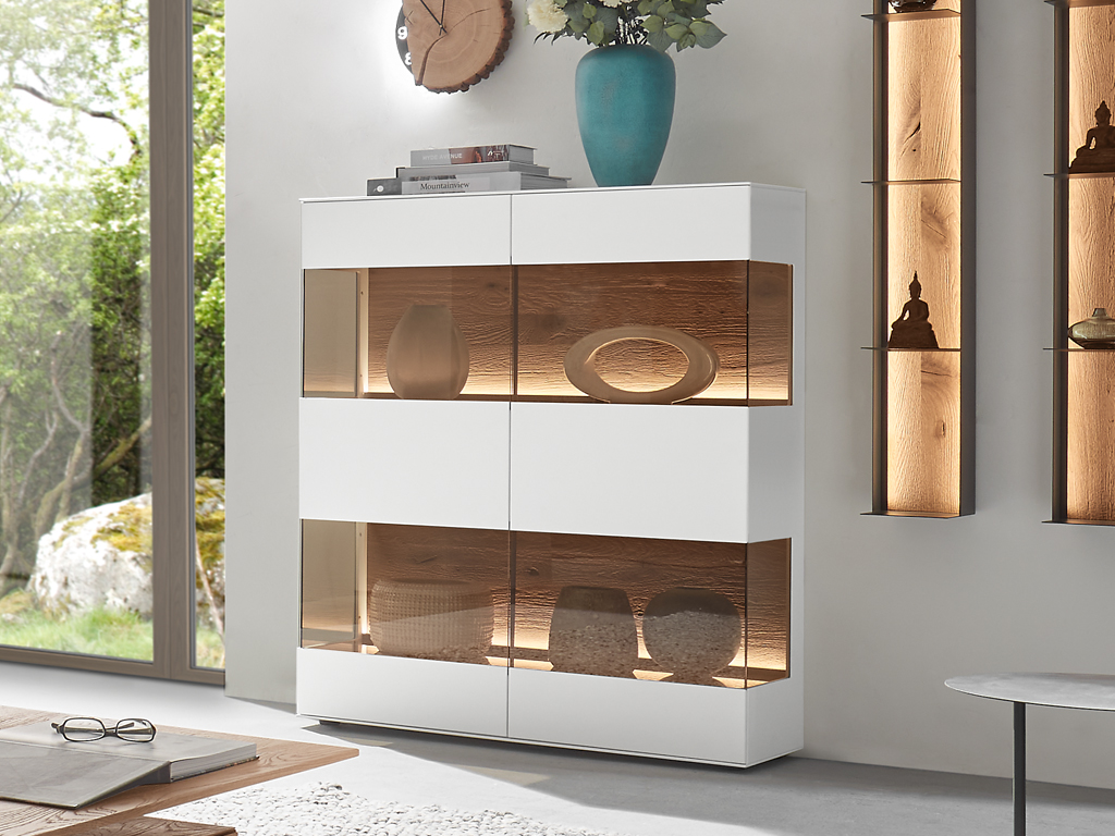 in modernes, weißes Hartmann Torido Highboard 7112 mit beleuchteten Holzfächern und Glastüren, das in einem hellen Wohnzimmer mit Blick ins Grüne steht. Die Fächer sind mit verschiedenen Dekorationsgegenständen bestückt.