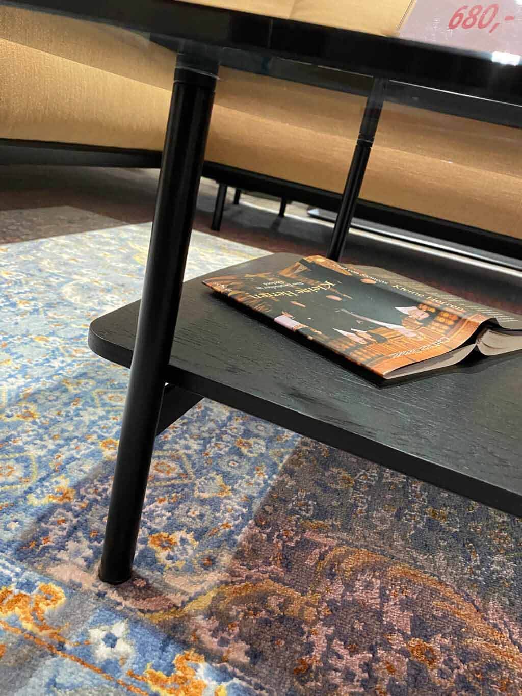 Nahaufnahme eines schwarzen Couchtisches mit Zeitschrift auf einem bunten, gemusterten Teppich, im Hintergrund ein helles Sofa sichtbar.
