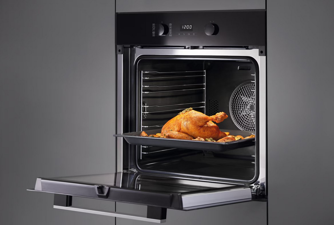 Ein geöffneter schwarzer Miele-Ofen, in dem ein goldbraun gebratener Hähnchen mit Beilagen auf einem Backblech auf einem Rost liegt. Der Ofen ist in eine moderne graue Küchenzeile eingebaut.