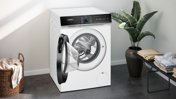 Eine Siemens iQ700 Waschmaschine mit geöffneter Tür steht in einem modernen Raum neben einem Wäschekorb und einem Beistelltisch mit gefalteter Wäsche.
