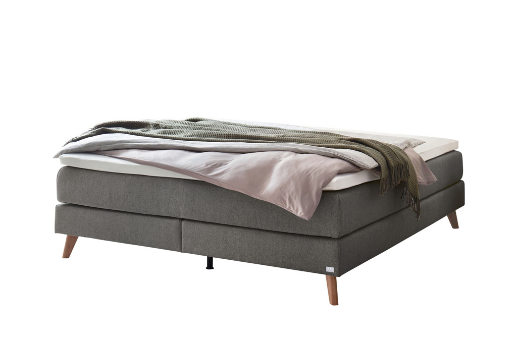 graues Boxspringbett mit braunen Holzfüße, stilvoll dekoriert mit Decken und Kissen vor weißem Hintergrund