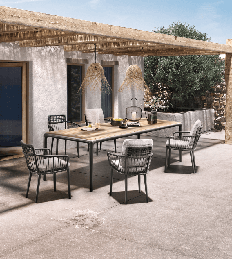 Outdoor-Esszimmer-Set mit Holz-Tisch, sechs Stühlen und Dekoration unter einer rustikalen Pergola neben einem Haus.