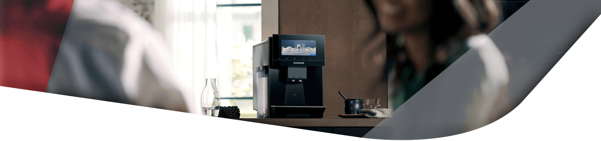 Ein schwarzer Siemens Kaffeevollautomat mit Touch-Display steht auf einer Küchenarbeitsplatte. Die Szene ist von unscharfen Personen im Vordergrund eingerahmt, wobei eine Person rechts angeschnitten zu sehen ist.