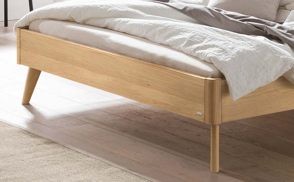 Detailaufnahme eines hellbraunen Holzbettes mit Holzfüßen in einem hellen Raum