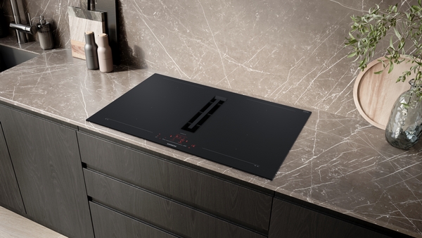 Ambienteaufnahme des schwarzen Siemens Induktionskochfelds mit integriertem Dunstabzug, eingelassen in eine helle, marmorierte Küchenarbeitsplatte über dunklen Holzschränken.