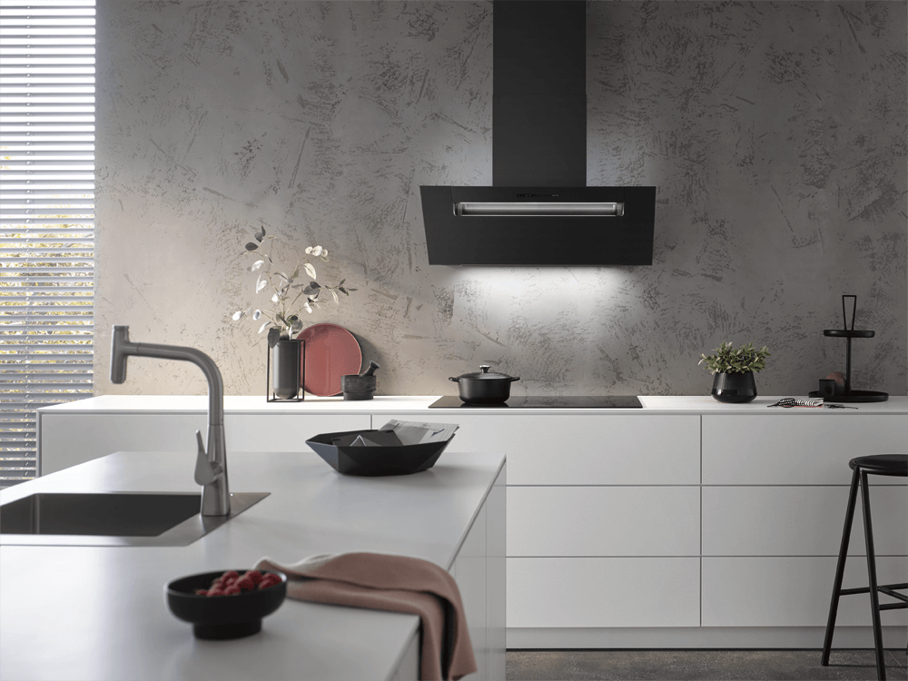 Eine stilvolle Küche hat eine mattschwarze schräge Dunstabzugshaube als Blickfang, die mit einer strukturierten grauen Wand kontrastiert. Die weißen Schränke und der helle Holzboden schaffen einen modernen und einladenden Raum mit natürlichem Licht.