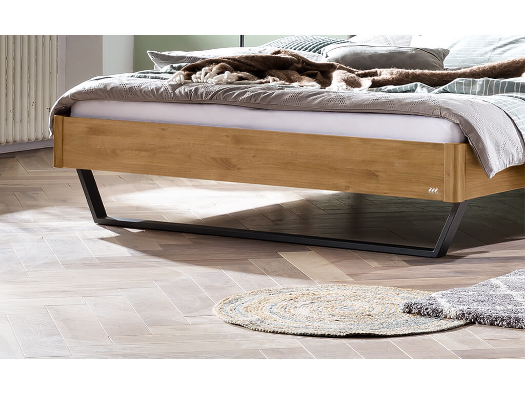 Nahaufnahme des Fußendes eines Bettes mit einem naturbelassenen, geölten Wildeichenrahmen. Das Bett steht auf einem schwarzen Metallgestell. Eine Bettdecke in Weiß mit einem Streifenmuster ist sichtbar.