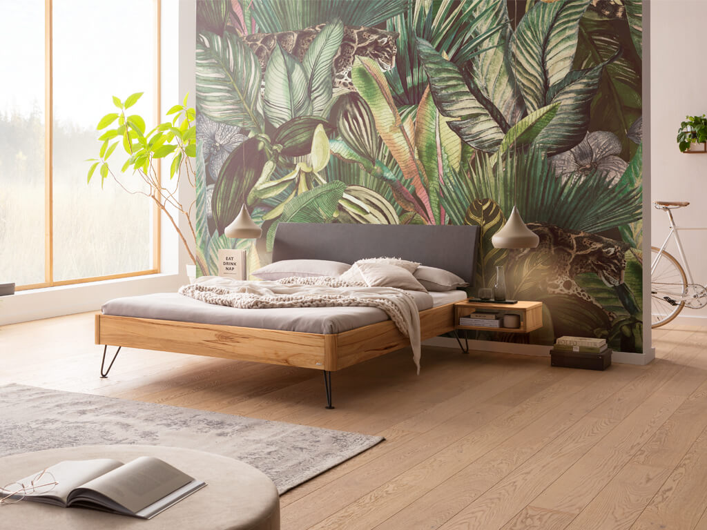 Ein Schlafzimmer mit einem Bettrahmen aus Wildbuche und einem anthrazitfarbenen Kopfteil. Die Nachtkästchen sind schwebend und ebenfalls aus Wildbuche. Die Wand hinter dem Bett hat eine Dschungel-Tapete und der Boden ist aus hellem Holz.