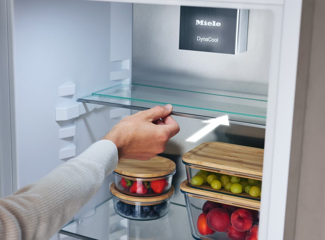 Eine Hand schiebt eine Glasablage in einem Miele Kühlschrank. Ein weißer Pfeil verdeutlicht die Möglichkeit, die Ablage zu bewegen. Auf den unteren Ablagen stehen Glasbehälter mit frischen Beeren und Trauben.