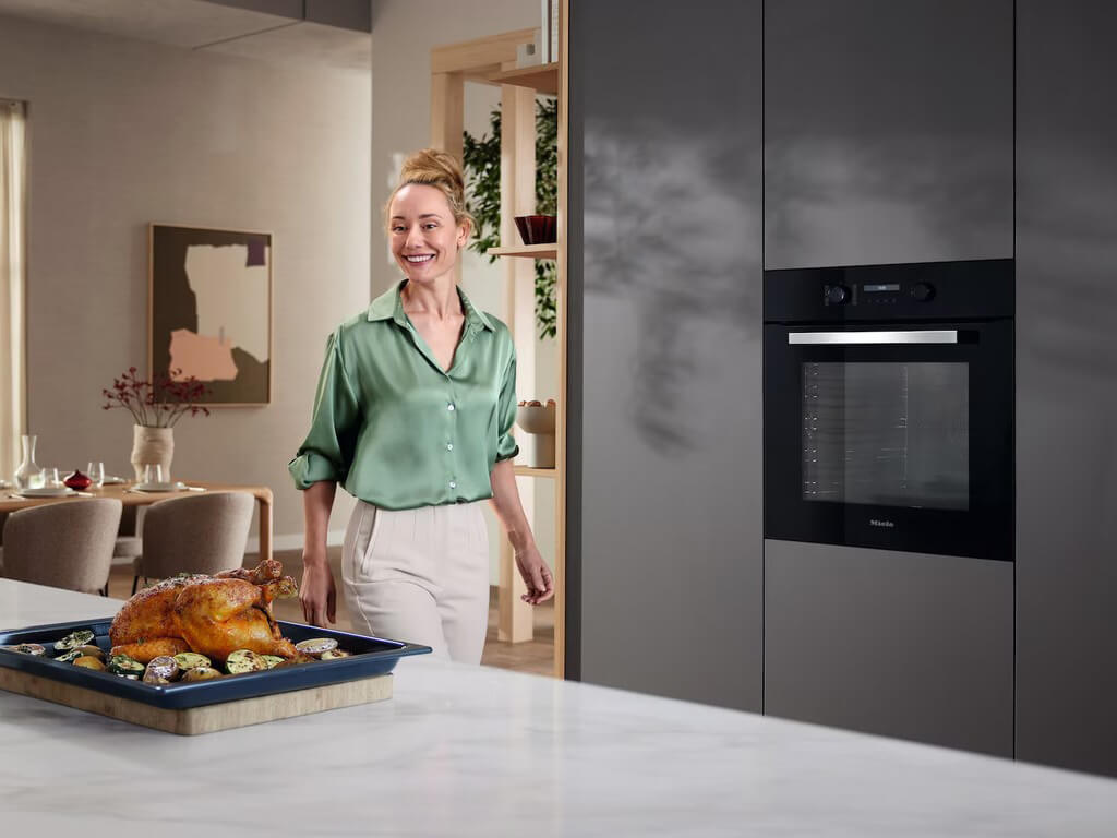 Eine lächelnde Frau in einem grünen Seidenhemd steht in einer modernen Küche neben einem Backofen.