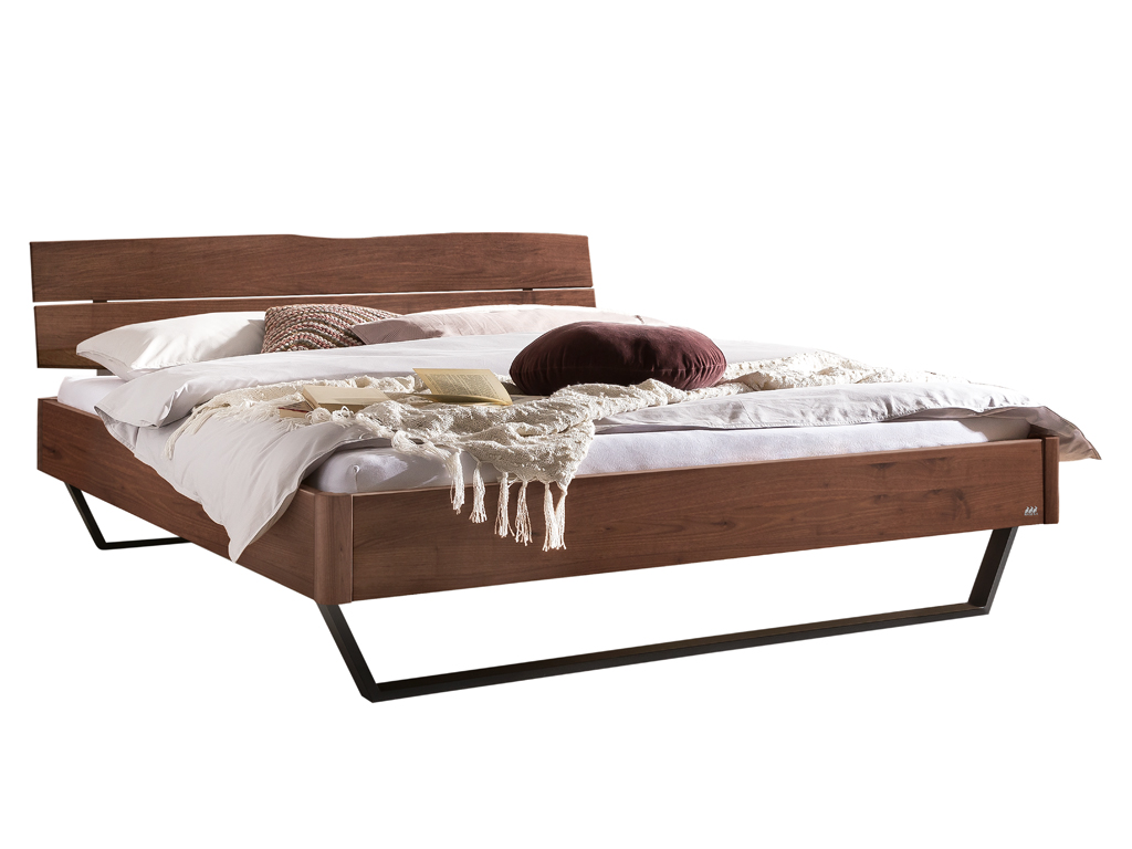 Ein freigestelltes Produktfoto eines Doppelbettes mit einem Bettgestell und Kopfteil aus dunklem Nussbaumholz. Das Bett steht auf anthrazitfarbenen Metallkufenfüßen und ist mit weißer und beiger Bettwäsche sowie Kissen dekoriert. Der Hintergrund ist weiß.