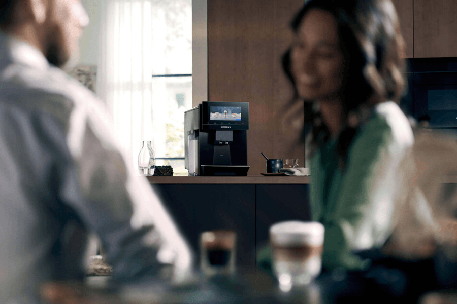 Nahaufnahme einer schwarzen Siemens Kaffeevollautomat auf einer Küchenarbeitsplatte, mit zwei unscharfen Personen im Vordergrund, die sich unterhalten.