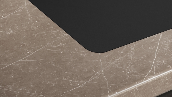 Detailaufnahme der abgerundeten Ecke eines schwarzen Induktionskochfelds, das bündig in eine braun-marmorierte Arbeitsplatte eingelassen ist.