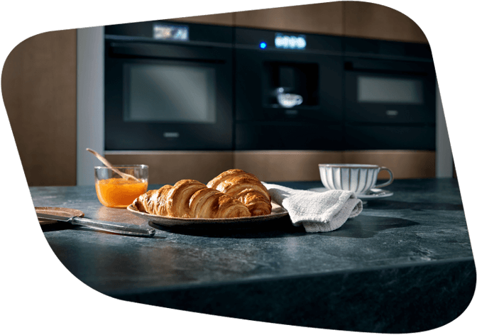 Ein Teller mit frischen Croissants, Marmelade und einer Tasse Kaffee auf einer Küchenarbeitsplatte, im Hintergrund moderne Küchengeräte