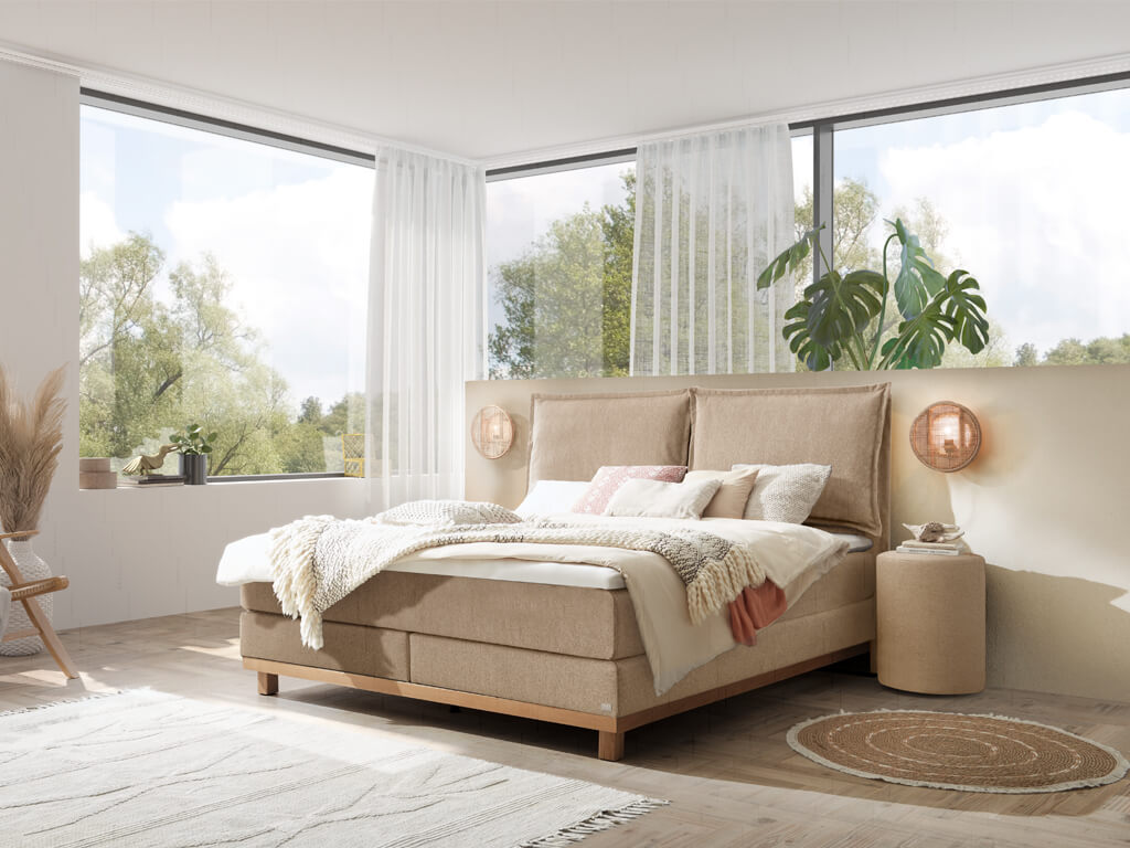 Boxspringbett in Sandfarben mit hellem Holzbettgestell und hoher, zweiteiliger Lehne steht in einem hellen Schlafzimmer mit großen Fenstern und Holzboden.