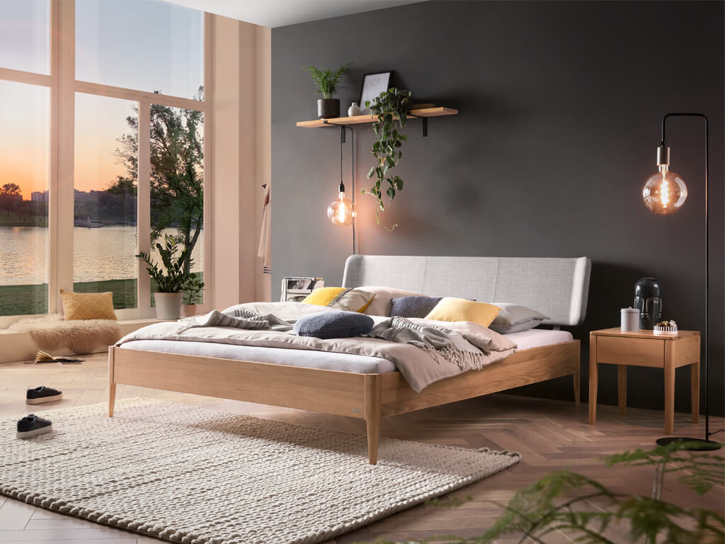 Ein gemütliches Schlafzimmer mit einem Hasena-Bett "Pienza Styly" aus heller Eiche "bright" und einem gepolsterten Kopfteil in "Pepe grey". Der Raum ist durch große Fenster mit Tageslicht erfüllt. Das Bett ist mit einer farbenfrohen Bettwäsche dekoriert.
