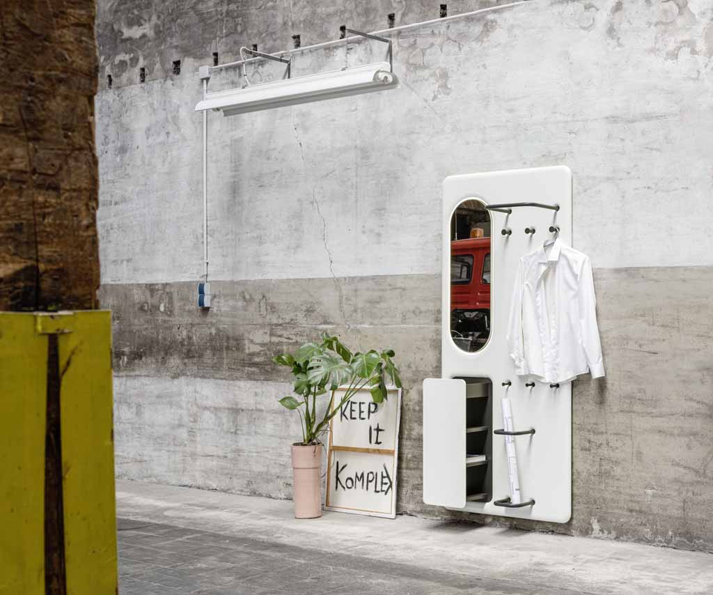 Sudbrock Juno -  Garderobenpaneel | B: 90,0 cm | Lack blütenweiß | Ausrichtung links Eine moderne weiße Garderobe mit Spiegel und Kleiderhaken, daneben eine Pflanze und ein Schild mit der Aufschrift 'Keep it Komple' in einem industriellen Raum.
