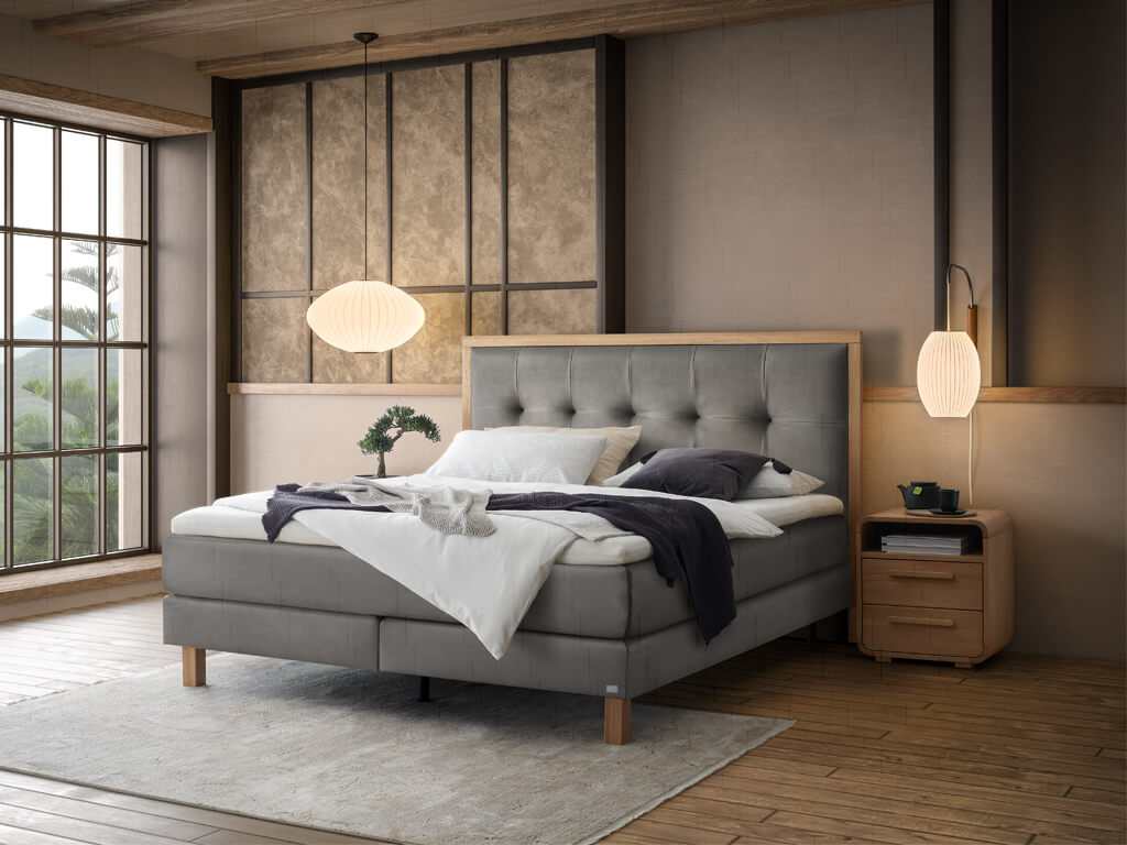 Ein Hasena Boxspringbett Pronto Solara Presley mit stahlgrauem Stoffbezug in einem Schlafzimmer, das durch dunkle Holzdetails und eine große Fensterfront geprägt ist.