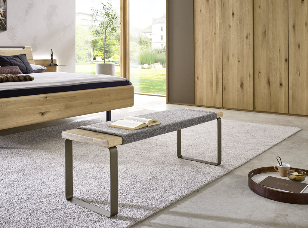 Moderne Sitzbank mit grauen Kufen und grauem Polsteraufsatz präsentiert vor einem Bett und stilvoll dekoriert in einem modernen Zimmer
