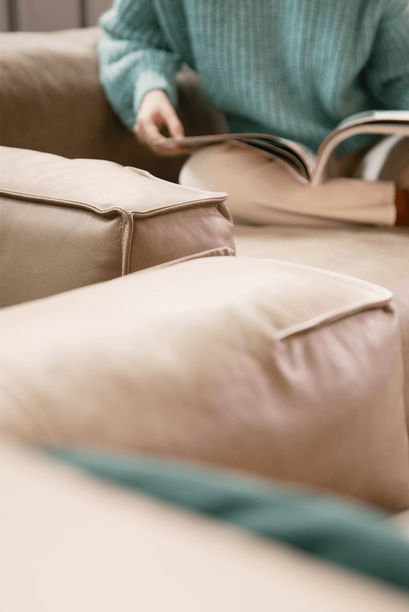 Person in einem blauen Pullover blättert in einem Magazin auf einem beigen Ledersofa