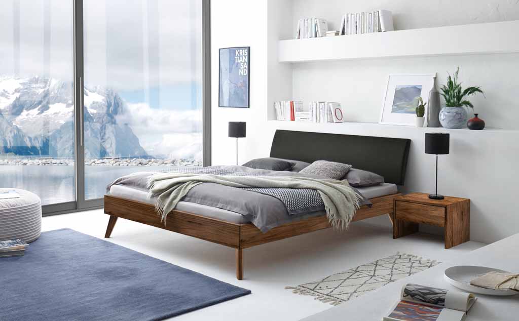 Helles, modernes Schlafzimmer mit einem Holzbett, flankiert von Nachttischen. Großes Panoramafenster mit Berg- und Seeblick, dekoriert mit Büchern und Pflanzen.