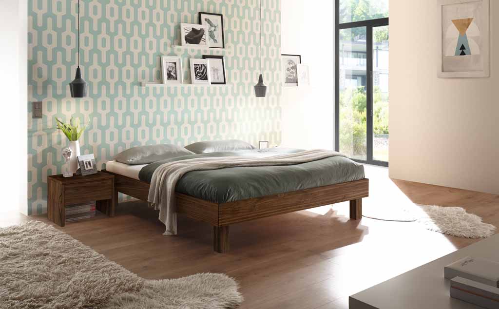 Ein gemütliches Schlafzimmer mit Holzbett, Nachttisch, dekorativer Tapete und großem Fenster mit viel Tageslicht.