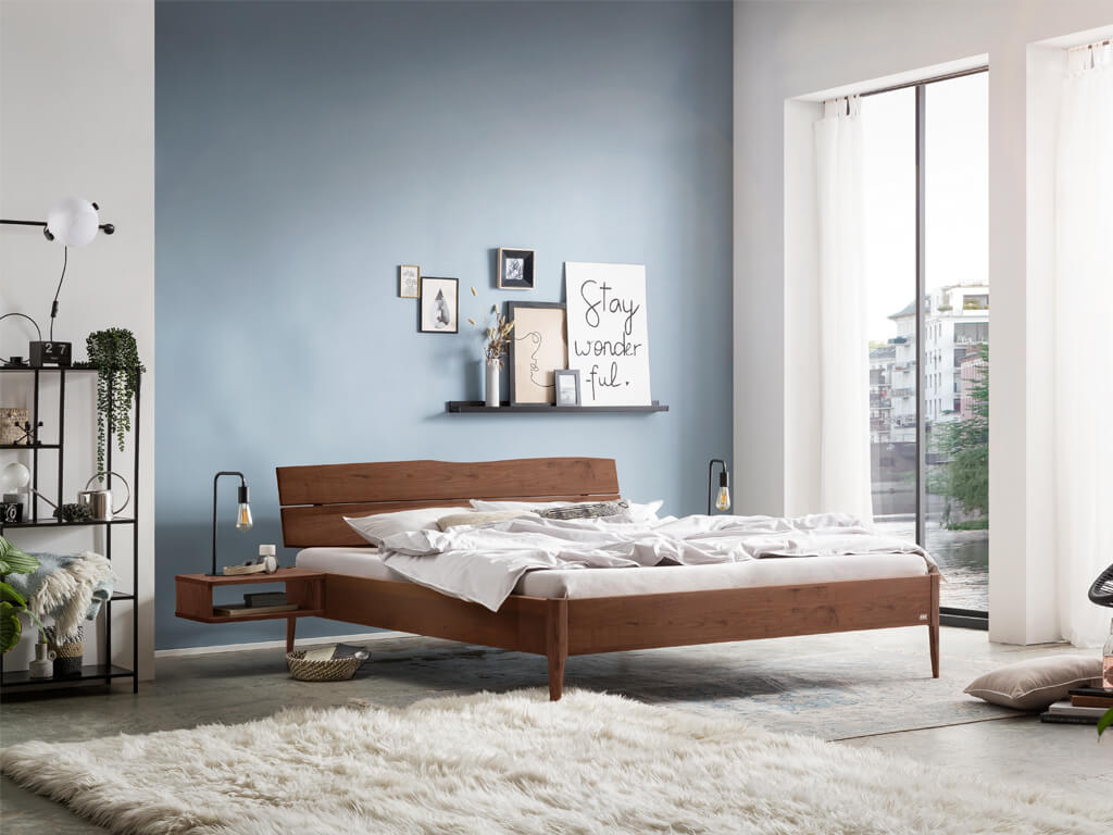 Hasena -  Lounge Bett Sabbioni | Holzkopfteil Turano | Holzfuß Pile | versch. Größen & Farben konfigurierbar dunkles Holzbett mit Kopfteil aus Holz in einem Schlafzimmer mit großem, flauschigen Teppich in weiß