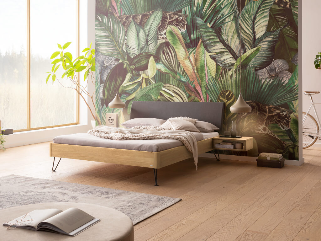 Ein Schlafzimmer mit einem Bettrahmen aus heller Eiche und einem anthrazitfarbenen Kopfteil. Die Nachtkästchen sind schwebend und ebenfalls aus heller Eiche. Die Wand hinter dem Bett hat eine Dschungel-Tapete und der Boden ist aus hellem Holz.