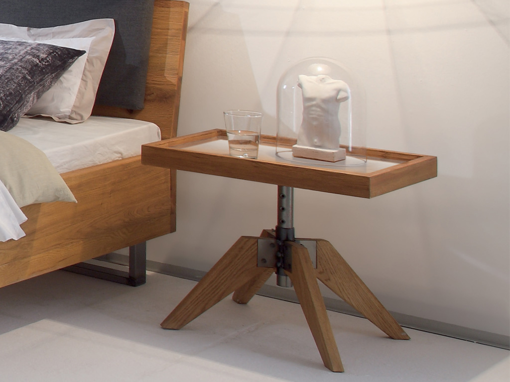 brauner exklusiver Holznachttisch auf dreibeinfüßen und einer Platte in Tablettform mit Glasablage stilvoll dekoriert an einem Bett Hasena Carpi 