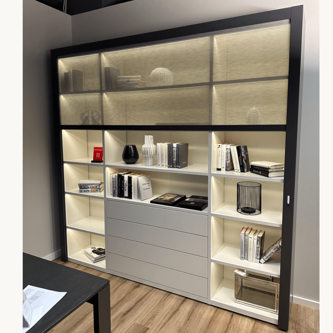 RMW COMFORT CLOSE | Wohnkombination mit textiler Front Ein modernes Regal mit integrierter Beleuchtung, dekorativen Objekten und Büchern, das in einem stilvoll eingerichteten Raum steht.