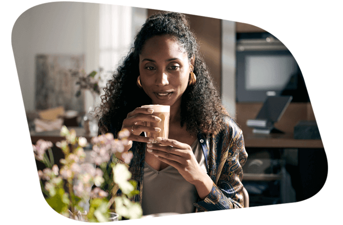 Eine lächelnde Frau mit lockigem Haar und goldenen Ohrringen hält eine Tasse mit einem Latte Macchiato oder Cappuccino. Sie sitzt drinnen an einem Tisch, im Hintergrund ist eine moderne Küche erkennbar.