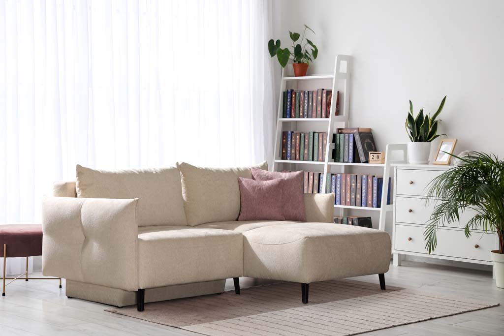 Restyl Landon - Schlafsofa (Sofaecke) | beige | verstellbare Armteile | 135 x 200 cm Helles Wohnzimmer mit beige Ecksofa, Bücherregal, Pflanzen und Kommode vor großen Fenstern
