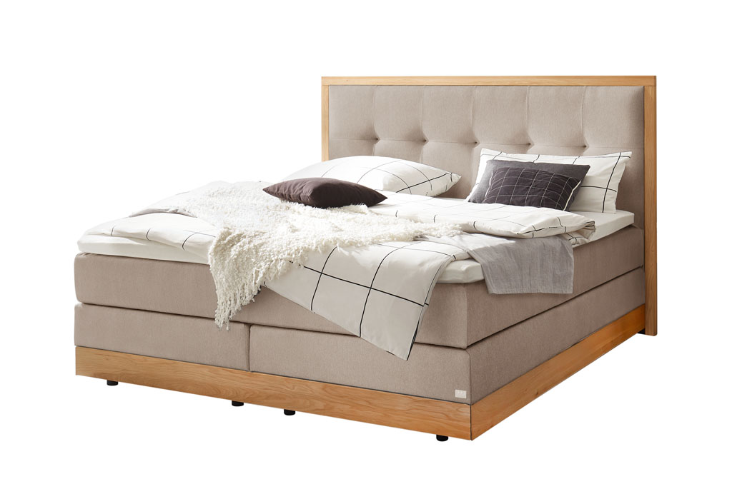 beiges Boxspringbett mit braunem Holzrahmen, stilvoll dekoriert mit Decken und Kissen vor weißem Hintergrund