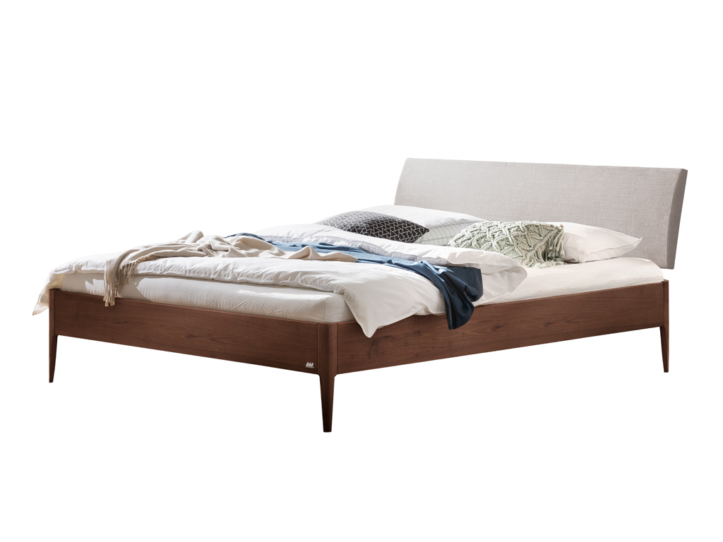Ein fertig montiertes Hasena Fanano Bett mit einem Bettrahmen aus dunklem Nussbaumholz und einem hellgrauen, gepolsterten Kopfteil. Das Bett ist mit weißen Laken und einer Decke sowie mehreren Kissen dekoriert. Der Hintergrund ist weiß.