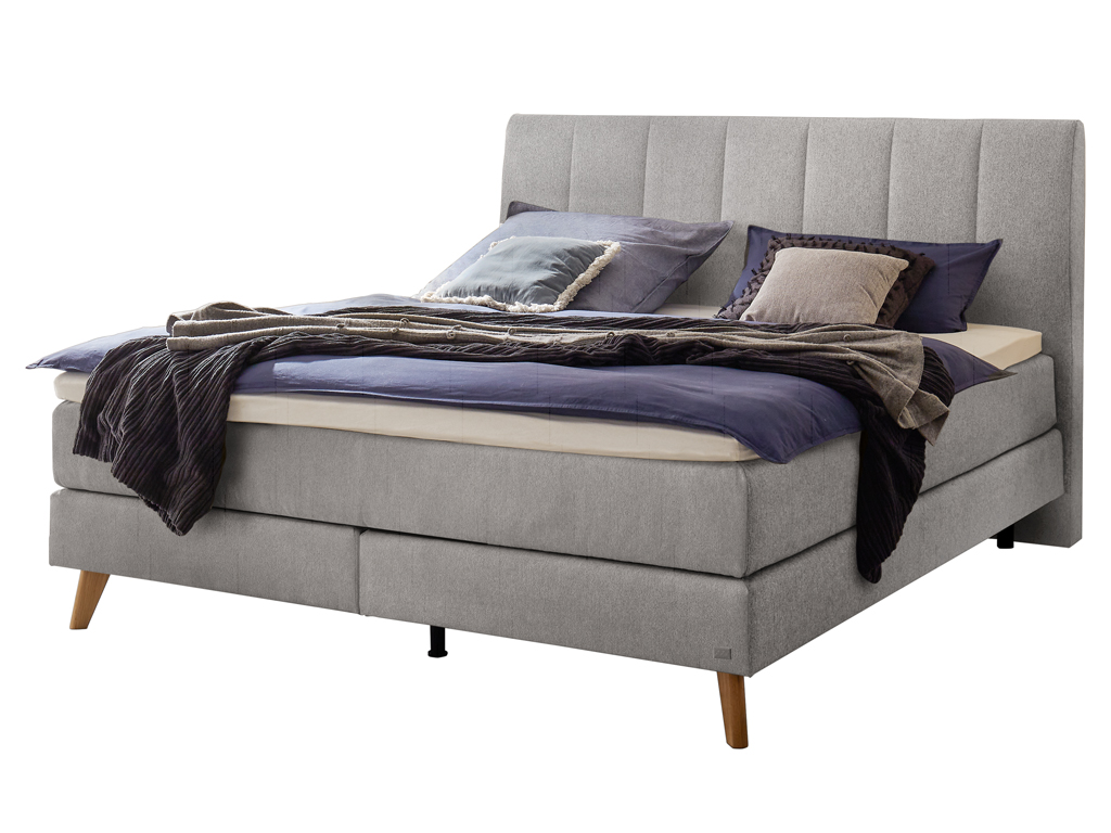 Ein Boxspringbett mit hellgrauem Kopfteil, Holzfüßen und passender Matratze und Bettwäsche vor einem weißen Hintergrund.