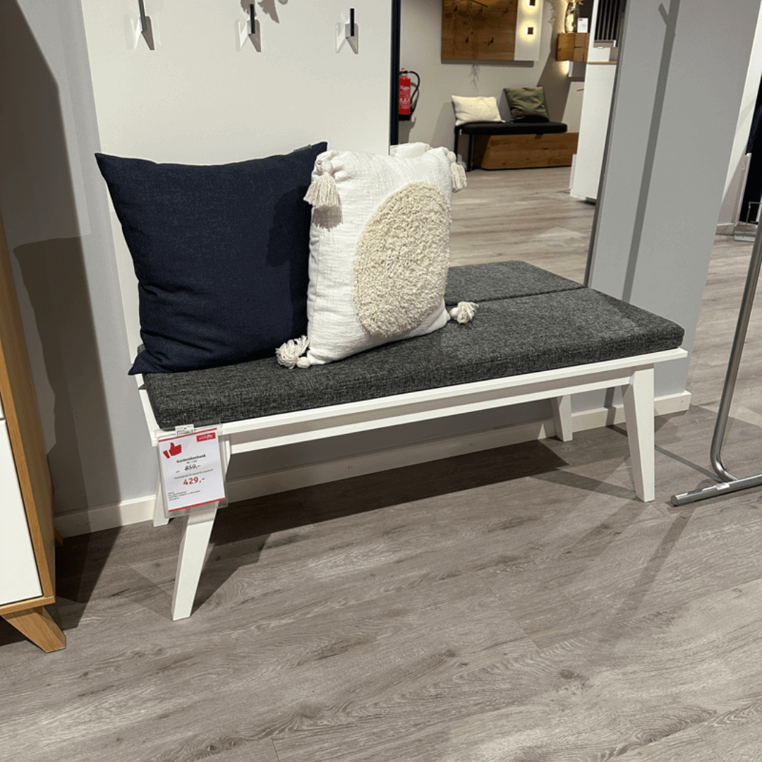 RMW DEVISO | Garderobenbank Weiße Sitzbank mit grauem Polster, zwei dekorativen Kissen, ausgestellt in einem Möbelgeschäft auf Laminatboden