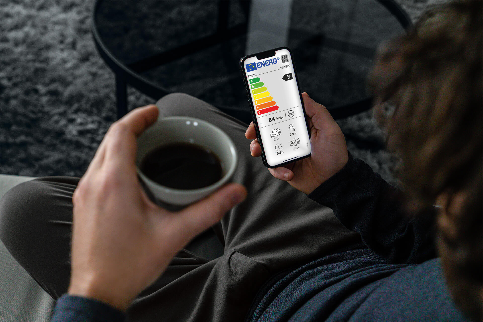 Person mit Kaffee und Smartphone in der Hand von oben fotografiert. Es ist ein Energieeffizienzlabel auf dem Smartphone zu sehen.