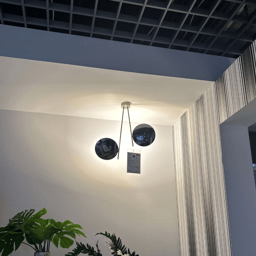 Stilvolle Deckenlampe mit zwei schwarzen runden Elementen, montiert an der Zimmerdecke. Im Vordergrund sind grüne Pflanzen sichtbar.