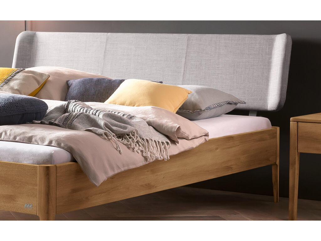 Eine Nahaufnahme des oberen Teils eines Bettes Pienza aus Wildeiche, natur geölt. Das Bett hat ein gepolstertes Kopfteil in hellem 'Pepe-grey' Stoff und ist mit verschiedenen Kissen in Beige, Grau und Gelb sowie einer gestrickten Decke dekoriert.