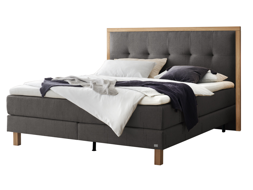 Ein freigestelltes Produktfoto des Boxspringbettes Hasena Pronto Solara mit dunkelgrauem Stoffbezug und Holzrahmen, dekoriert mit Decken und Kissen.